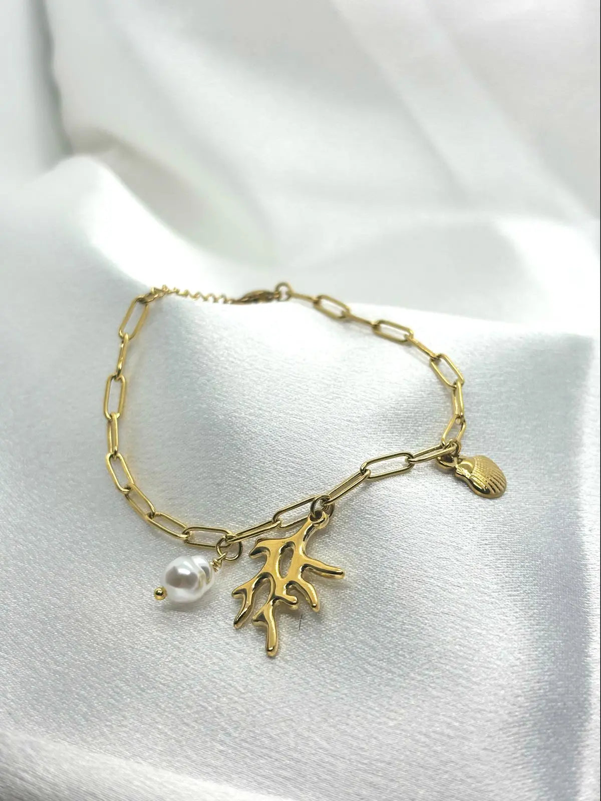 Bracelet doré avec breloques perle, corail et coquillage sur tissu satiné blanc.
